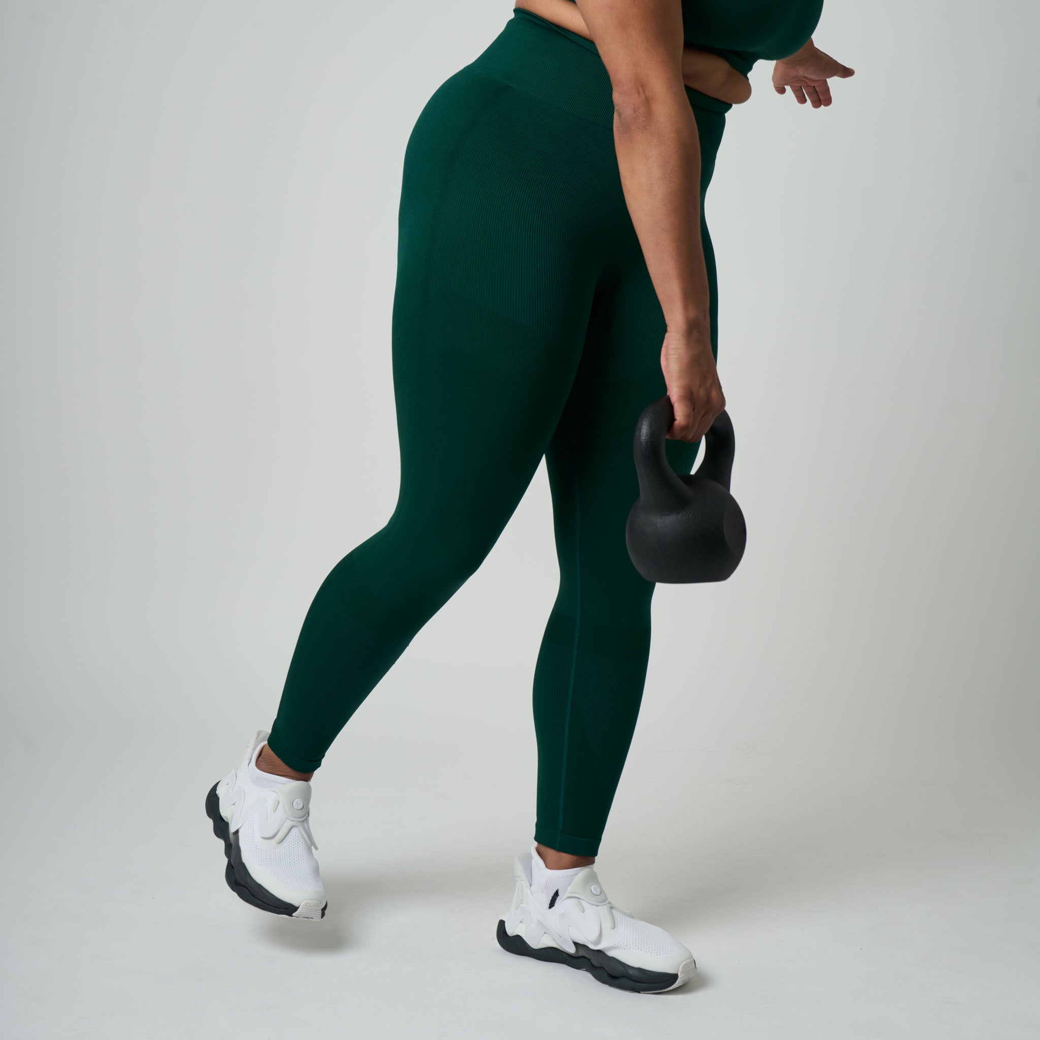Person wearing green leggings and white sneakers holding a black kettlebell on a light gray background