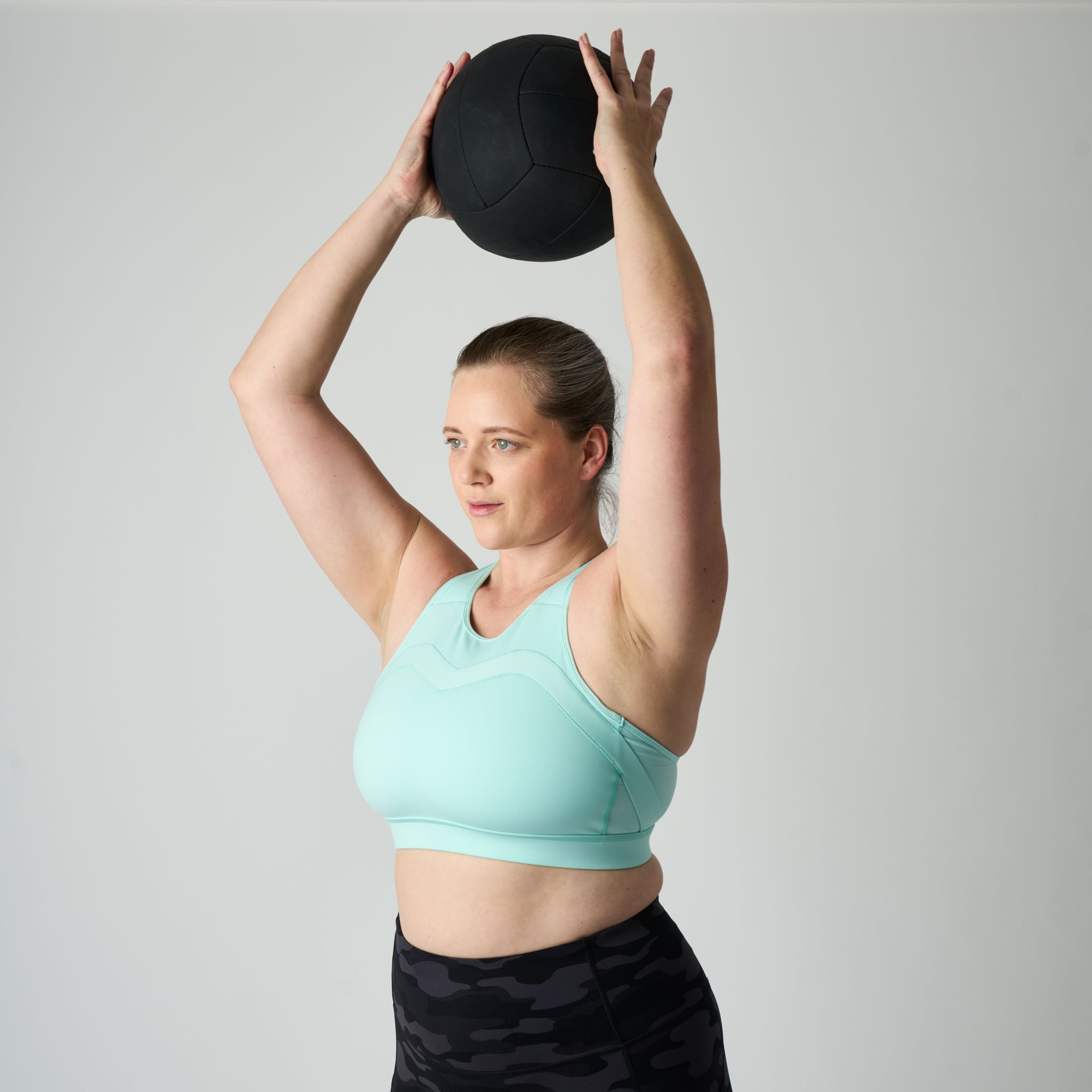 Woman in a light blue sports bra and black leggings lifting a medicine ball above her head against a gray background
